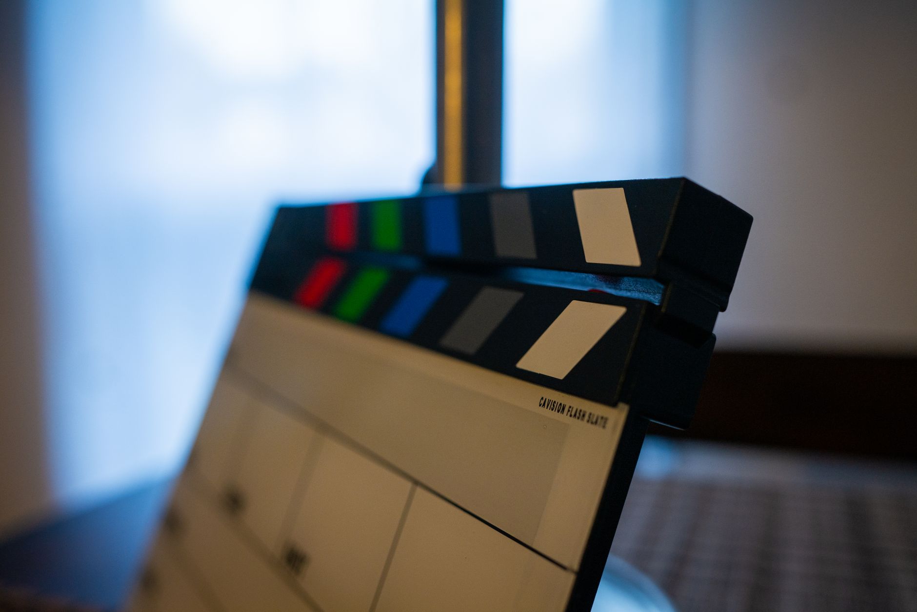 Gros plan d’un clap de cinéma noir et blanc aux bandes colorées, posé en intérieur devant une fenêtre lumineuse et floue.
