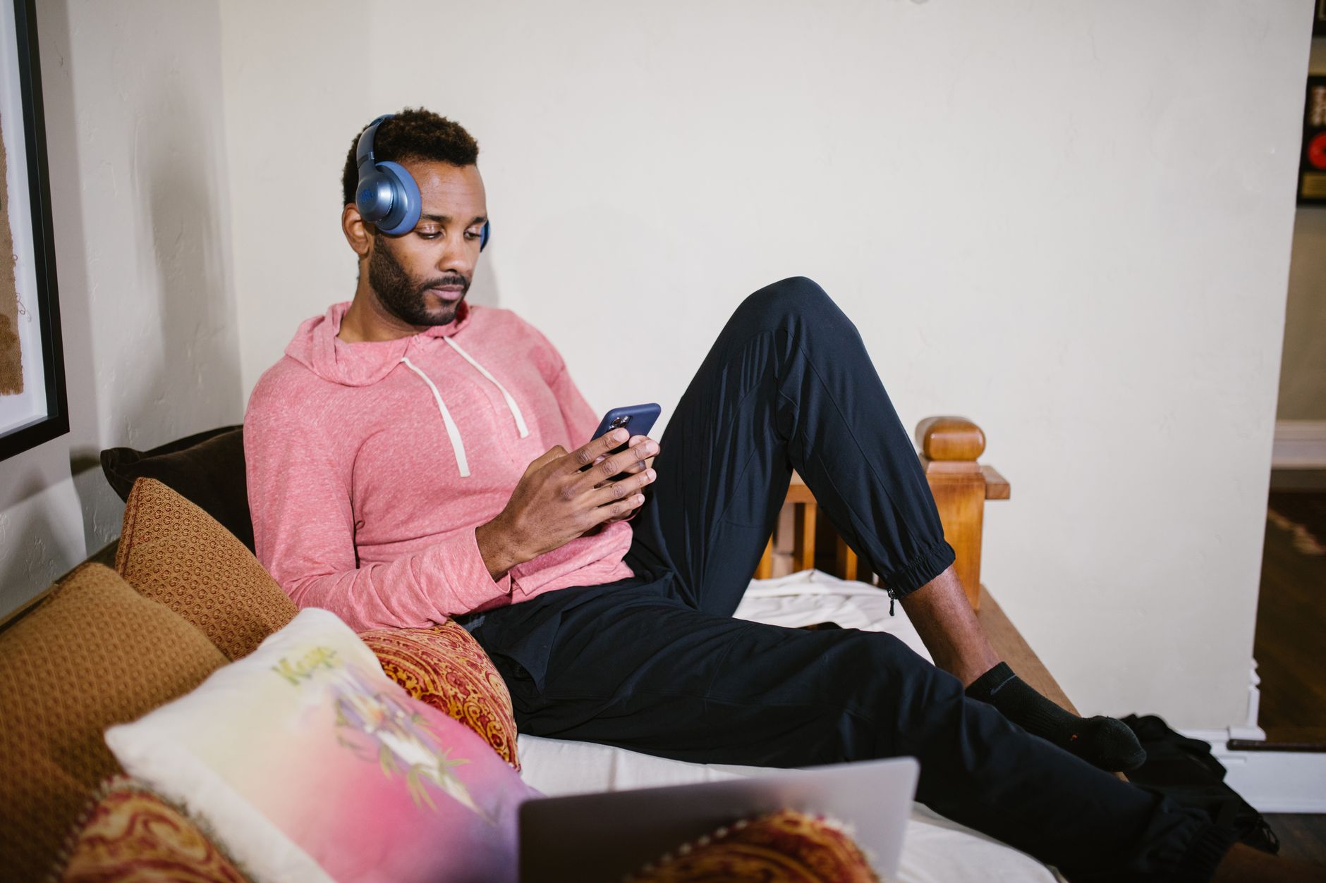 Homme en sweat rose, casque audio bleu sur les oreilles, regardant son smartphone sur un canapé garni de coussins.